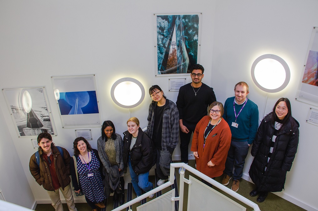 A group of 9 people standing in a white stairwell with framed photographs surrounding them